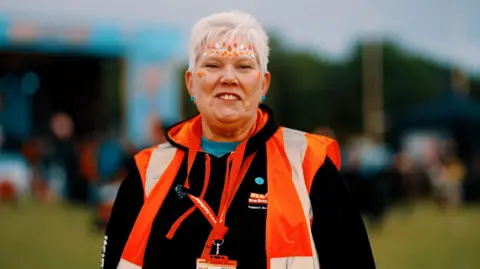 An older woman with short, white hair wears orange face gems and smiles as she volunteers at a festival. She is wearing an orange hi-vis vest, a dark hoodie and an orange lanyard.