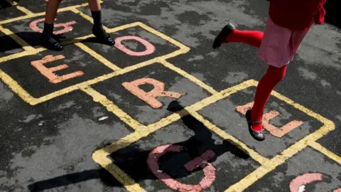 A stock photo shows two children playing hopscotch in a school playground.