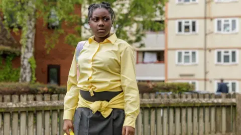 Girl with braided hair who is wearing a yellow shirt and grey skirt.