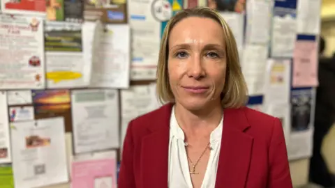 Helen Morgan - a woman with long blonde hair, wearing a red jacket over a white top - stands in front of a noticeboard and smiles.