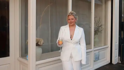 Georgia Sullivan Georgia smiling outside her cream salon. She wears a white two-piece blazer suit set and holds a glass of prosseco. She has blonde hair which is in a top bun. 