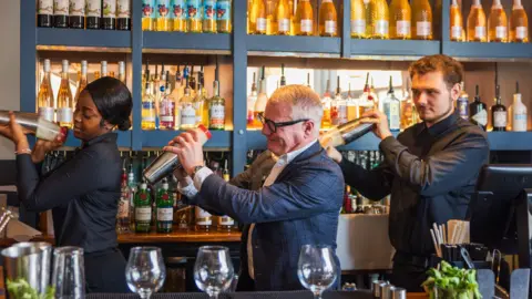 WMCA Two men and a woman stand in a line behind a bar, all shaking cocktail mixers. They are in front of blue shelving containing rows of bottles.
