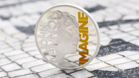 Royal Mint A silver coin with a portrait of John Lennon on it, with lettering that reads John Lennon on one side, and the words 'Imagine' in yellow, on the opposite side