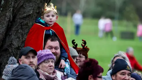 Reuters A boy wearing a plastic gold crown and red cape sits on the shoulders of a man with a crowd of people gathering ahead of the Christmas Day morning church service attended by the royal family at St Mary Magdalene Church in Sandringham, Norfolk. People are wearing wool hats and and chat among themselves.