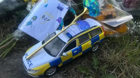 A yellow, blue and white toy police car is set among the floral tributes. There is a plastic flower set on top of it as well as a handwritten note. There is a drawing of police officers and the word "RIP" on a piece of paper wrapped around another bouquet of flowers on the left.