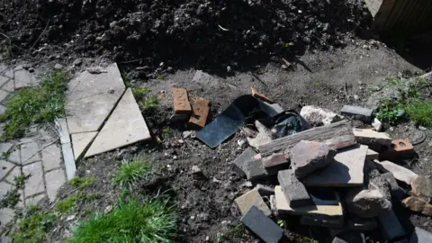 A patch of ground is covered with broken bricks, concrete pieces and other building debris. The rubble sits on soil and paving stones, showing an unfinished or abandoned construction job.