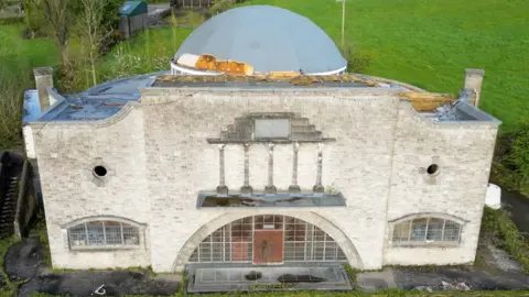 Owen McNutt Edendork Hall taken before its demolition with a light brick exterior and curved large windows, at the top of the building is an impressive large dome roof which is light grey with specks of gold (it would have been gold all over before deterioration. It stands on a plot of land with green grass behind it. 