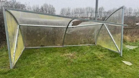 Stroud Times A broken transparent plastic shelter with a metal frame stands on grass, its roof collapsed and a side panel bent inward. A wire fence and bare trees are visible in the background beneath a grey, overcast sky.