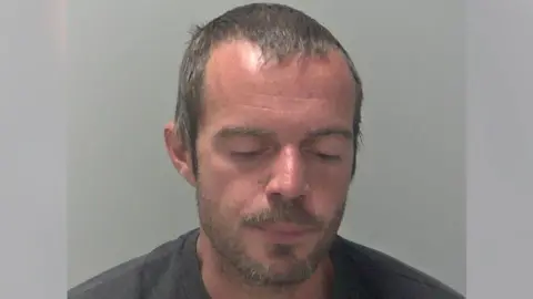 A man, Daniel Gibbs, looks down at the ground in a police mugshot. He has light brown, short hair and light brown stubble. He is wearing a dark-coloured top.