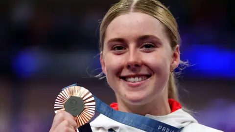 A female cyclist smiles as she holds up an Olympic bronze medal attached to a blue ribbon, which she is wearing around her neck. She has blond hair, tied back, and is wearing a white tracksuit top with red trim.