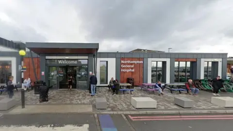 Google Hospital entrance with "welcome" in white lettering above the glass doors, and "Northampton General Hospital" picked out in white letters on a wooden panel to the right of the main doors. The doors have a grey portico in front of them. Grey block paving is visible in front of the building, and there are double red lines on the edge of a roadway in the foreground. Several people are either walking or sitting in the image.