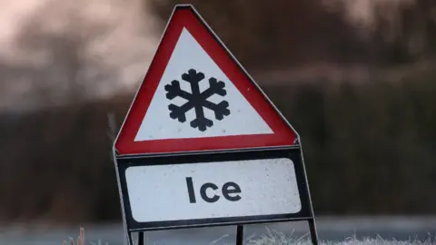 A road sign warning of ice,  The sides of the road are covering in snow. 