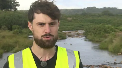 Lewis Edwards is wearing a yellow high vis jacket. He is standing in front of water.