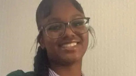 A close-up photo of a smiling Elianne Andam, she has long black hair in a braid and is wearing glasses. She is wearing a striped school uniform blouse.