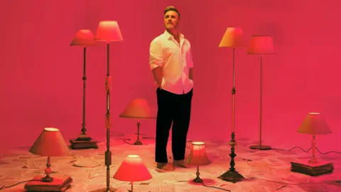 Gary Barlow, wearing a white shirt and black trousers, stands beside a number of upright lamps in front of a red-coloured background   