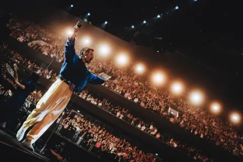 Netflix Harry Styles on stage in Manchester's Co-Op Live Arena, wearing baggy cream pantalons and a navy blue shirt. He has his hands in the air, while thousands of fans watch from three levels of seating and lights shine on the stage,