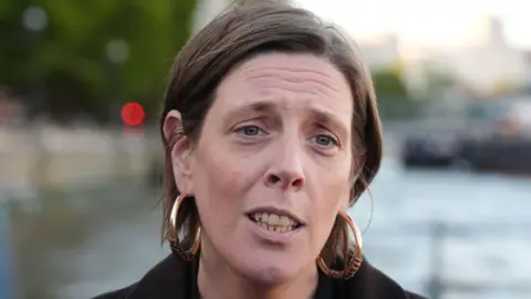 Jess Phillip is outside. She has short brown hair. She's wearing gold hoop earrings. The background is out of focus.