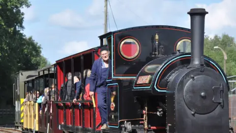 A black steam engine can be seen pulling a series of carriages. A man wearing navy blue overalls is leaning out of the cab. There are a number of passengers in the open-sided carriages behind.