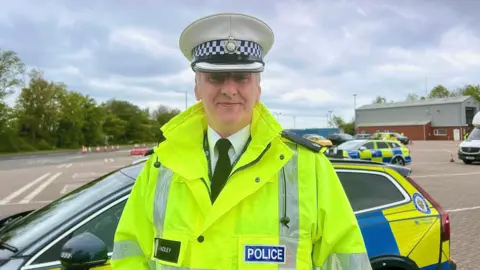 Jack Hadley is smiling into the camera. He is wearing a neon yellow/green high vis jacket and it says POLICE. He is also wearing a police hat which is white. There is a car behind him, which is a blue and yellow police car. 