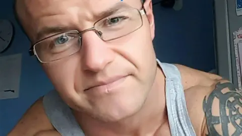 Social media A man in close up looking into the camera, He is wearing a grey vest and his bare shoulders are apparent, a tattoo with a spiral pattern is visible on his left arm. He is wearing glasses and appears to have closely shaved hair. He is inside, the background is a blue wall and a clock is partially visible. 