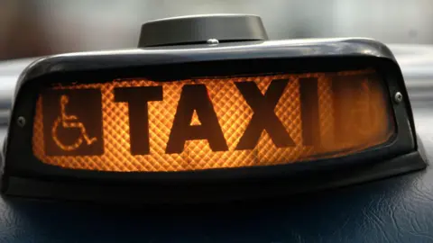 An illuminated taxi sign on top a vehicle. It has a disability sign, depicting a person in a wheelchair, to the left of the sign.