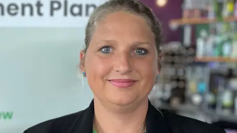 Victoria Oliver - She has blonde hair tied back behind her head, is smiling at the camera and has blue eyes. She's wearing a black suit and a green blouse. She is standing in front of a sign about the focussed review of the Island Development Plan.