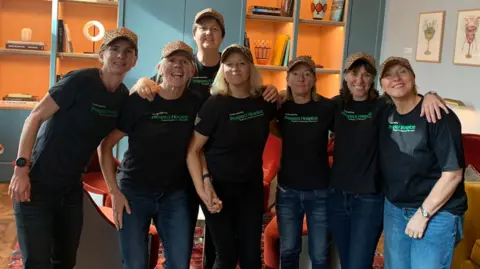 Prospect Hospice The five women and two of their support team, in a line-up, smiling to camera, wearing black t-shirts with the Prospect Hospice logo on the front and leopard print baseball caps, standing in a large hotel-type room 