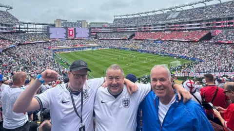 Paul Dubberley Three football fans posing with a packed out football stadium behind them at an England match