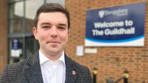 A dark-hair man standing in front of brick building, wearing a navy blazer, white shirt and small red poppy on his lapel. A sign in the background reads, "Welcome to the Guildhall".