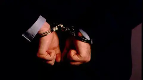 Close up of a man's handcuffed hands behind his back. He is wearing a black blazer with a light grey shirt underneath.