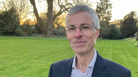 Sam Read/BBC James Talbot, who has short grey hair. He is wearing metal-framed glasses and a blue jacket over an open necked church. Behind him is lawn and beyond that a London plane tree.