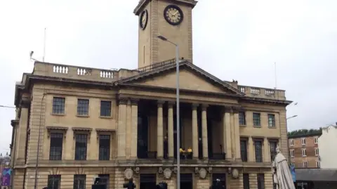 A large neoclassical civic building with a pale stone façade, tall columns forming a portico, and a central clock tower rising above the roof. A statue stands to the right of the building, with streetlights in front and an overcast sky in the background.