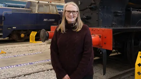 Sarah Price has long blonde hair and glasses and is smiling and wearing a long maroon jumper. She is standing in front of a black and red locomotive and a blue locomotive inside a museum.