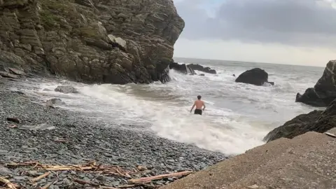 Shaun Kelly There is a pebble beach on the left and the sea is rough with white horses crashing onto the stones. Shaun Kelly is walking into the sea in a small cove with a sharp cliff side to the left and jagged rocks on the right - it is a grey day.