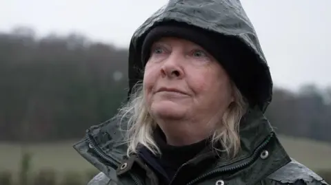 Deborah wears a hat and raincoat. She is outside, looking off camera, and looks determined.