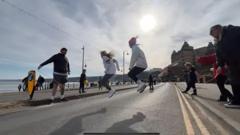 Oli Constable/BBC A man and a woman hold either ends of a skipping road. Two girls are jumping over it. They are standing on the road on the Scarborough foreshore. 