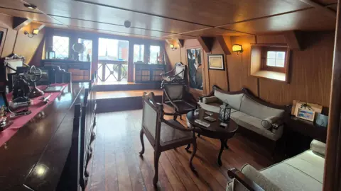 A warmly furnished, wood-paneled interior of is the admiral's quarters on a historic ship, with several chairs and a table.