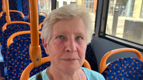 John Devine/BBC Patricia Chalkley, 76, has light blonde short hair with a turquoise V-necked jumper on with a gold chain around her neck. She is sitting on the blue and orange seat of a bus with a shopping bag beside her and empty seats behind her. 
