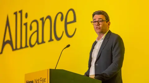 Pacemaker Andy Burnham is standing behind a podium and speaking into a microphone. He is wearing a dark grey suit with a white shirt underneath. His collar is open, with no tie. He is wearing dark framed glasses. Behind him is a yellow wall with the word Alliance written on it. 