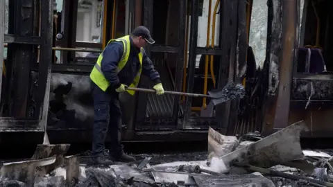 PA Media A man in a navy overall wears a hi-viz vest as he shovels debris away from the remains of a burnt out bus. The bus has been charred black. 
