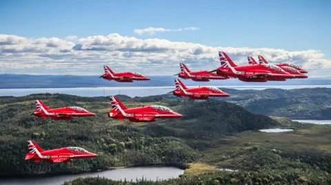 MOD Red Arrows team flying over a vast Canadian landscape, carpeted with greenery. The team are in formation.