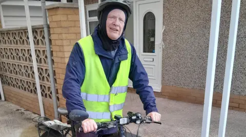 Tudur Davies standing outside a house and holding a bike. He is wearing a blue coat with a high vis vest over the top, along with a balaclava and a bike helmet