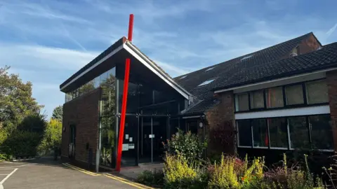 A modern or renovated church building, with a glass frontage, and a distinctive red column outside the entrance. The photo is taken from a car park area. There is foliage in the foreground and it is a sunny day.