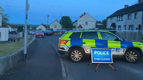 A police cordon in the area. There is a police car, tape and a road closed sign
