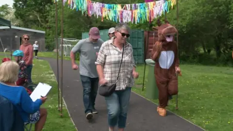 A group of people at an outdoor event on a grassy area with trees and playground equipment in the background. Colourful streamers hang above a pathway where individuals are walking or jogging. One person is dressed in a brown cow costume. Others nearby are standing or sitting, some holding clipboards, appearing to organise or monitor the event.