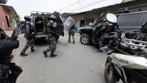 Polícia Equadoria em Protetive Gar e carregando armas montadas em uma estrada do lado de fora de um prédio, cercado por veículos policiais