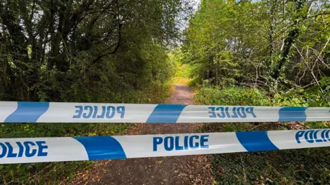 police tape in blue and white over a footpath in Wakefield with trees on either side 