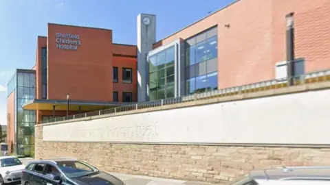 The exterior of a large, modern hospital building. The main structure is made of red brick with sections of glass and metal, giving it a contemporary appearance. On the left side of the building, there is a prominent sign that reads “Sheffield Children’s Hospital”.