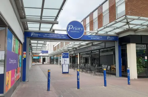 Local Democracy Reporting Service The entrance to the existing centre with a row of blue bollards on a pedestrianised route and a big blue sign above