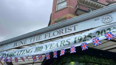 A close up shot of the June the Florist shop sign, a banner reads "celebrating 100 years 1025-2025". There is Union Jack bunting below the banner.